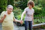 Aging Alzheimer’s patient with short white hair and wearing glasses in a yellow blouse holding hands with a middle-aged woman with curly brown hair