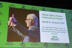 A large video screen above a stage shows David Amaral doing a thumbs up sign with text that reads INSAR Lifetime Achievement Award 2026,UC Davis distinguished professor of psychiatry and behavioral sciences and MIND Institute with the INSAR logo