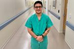 Medical student with dark hair, eyeglasses, green scrubs and tattoo on his left arm stands with hands crossed in an empty hallway at UC Davis Medical 