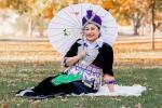 Smiling woman in traditional Hmong clothing sits on a blanket over grass. She is holding a parasol decorated with floral designs