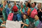 Thirteen health care providers in scrubs pose for a photo with a bunch of backpacks in a hospital room.