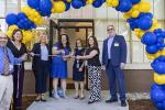 A group of people gather under a blue and gold balloon arch for a ribbon cutting