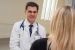 Martin Cadeiras with stethoscope around his neck sitting and speaking to patient with back turned. 