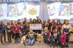 Twenty-four people and three dogs gather in a room holding an oversized check with $90,600 written on it