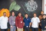 A group of seven people standing together with a “Central Kitchen” sign on the wall behind them.