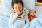 Girl lies in bed coughing, with her hand covering her mouth. A teddy bear is next to her.