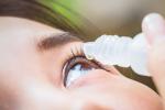 A woman holds an eye drop vial with a single drop above her eyes against a blurred background. 