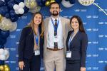 Three medical students – two women and a man – stand next to blue, white and gold balloons and a blue “School of Medicine” backdrop