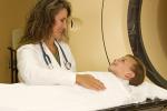 Child lays on gurney covered by white blanket as woman in white lab coat stands over him near circular CT machine. 