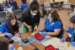 UC Davis Health surgeon Anamaria Robles leans over a table to show four teenage girls in blue polo shirts how to suture a banana