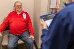 An older adult sits in a medical exam room chair while a healthcare worker stands nearby reviewing information on a tablet, with clinical equipment an