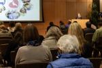 A crowd of seated people look toward a podium where a woman gives a lecture in an auditorium.