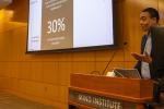 A man stands behind a wooden podium and in front of a wood-paneled wall. to his right is a large screen with a PowerPoint presentation projected