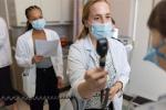 Student wearing mask and white coat extends arm holding ophthalmoscope up to patient’s eyes while other students look on