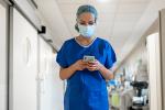 A masked female physician wearing blue scrubs looks down at a smartphone while walking down a hospital corridor.