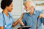 A woman in blue scrubs and holding a tablet sits on a couch next to an older, white-haired man resting is hand on a wooden cane. 