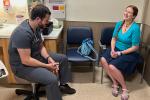 A man with dark hair and beard, in gray scrubs, talks with a woman with long braided hair and blue top in a clinic exam room 