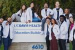 A group of 13 medical students wearing white coats next to the “Education Building” sign on the UC Davis Health campus