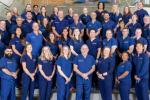 Dozens of emergency medicine team members in matching blue scrubs stand on a staircase inside a modern building.