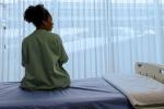 A woman sits on a hospital bed in front of a large window. 