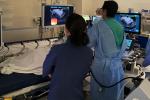 Three people wearing surgical scrubs standing next to patient lying in bed looking at ultrasound monitors