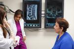 Two medical providers in white coats chat with a patient wearing a bright blue shirt in a medical exam room. 