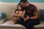 A father and daughter sit on a couch, the child’s knees are up at her chest, and the father is leaning down, consoling her, with his arm around her. 