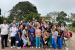 A large group of healthcare professionals wearing scrubs and badges pose outdoors, with trees and buildings in the background.