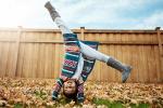 A young girl with long dark hair, wearing a bright multicolored sweater and gray boots does a handstand outside in a yard covered with leaves, in front of a wooden fence. 