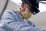 Surgeon wearing blue scrubs with bright lights behind him leans over patient wearing large black headset.