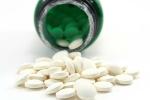 Round, off-white folic acid pills spill out from the mouth of a green vitamin bottle against a white 