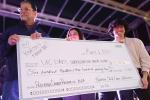 Three people on stage, man with dark hair accepts giant check from woman and young man.