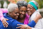 Three older smiling African American women in an outdoor park embrace in a group hug.