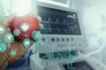 A hand holds a model of a heart in front of medical device’s digital screen