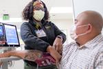 Nurse in gray fleece sweater and a mask over her face takes the blood pressure of a patient wearing a mask over his face.