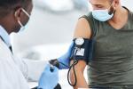 A doctor in a white coat and surgical mask holds a gloved hand on a blood pressure cuff that’s wrapped around the left arm of a young man