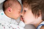 A young boy touches his nose to his infant sibling’s nose as the two cuddle in a bed.