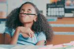 A young girl sits at a school desk, balancing a pencil between her nose and her lip while looking up at the ceiling. 