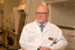 A man with blondish hair, mustache and beard wearing glasses and a white lab coat sits in a research lab facility.