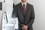 A brown-haired man in a gray suit with cream-colored shirt and burgundy tie stands next to an MRI machine. 