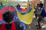 Children on a playground laugh while tossing a brightly colored parachute in the air. 