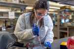 A person in a lab setting is carefully adding liquid to a lab tray using a dropper while wearing safety gear.