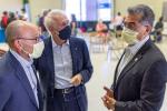 Three adult men wearing face masks gather in conversation while patients get vaccinations in the background.