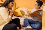 A woman and a young boy sit on the floor in a warm, inviting classroom, smiling and playing wooden string instruments together.