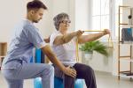 Male employee helps female senior patient with a physiotherapy exercise on fit ball. Woman on fit ball exercises with rubber band.
