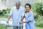 An older man using a mobility walker outside is assisted by a nurse. 