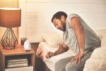 A man wearing gray sweatpants and a gray t-shirt sits with feet on the floor next to the bed and eyes closed. His nightstand holds an alarm clock and lamp. 