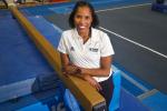 A woman wearing a white polo shirt and watch with a red band stands in a gymnasium leaning on a brown gymnastics balance beam and smiling. 