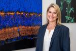 Smiling woman in blazer stands beside colorful microscopic image artwork displayed on a wall.