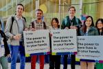 Seven medical students stand in front a building while holding signs encouraging signups for the National Marrow Donor Program